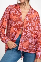 Blusa oversize de poliéster con estampado de animales y plantas en tonos rosa y marrón. Tiene cuello camisero, mangas largas y cierre frontal con lazos.