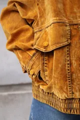 Campera estilo bomber o trucker de color mostaza/naranja quemado con efecto acid wash o desgastado. Presenta cuello camisero, cierre frontal y puños y cintura ajustados con elástico. Posee bolsillos frontales con solapa y botón.