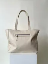 Cartera tipo tote color beige, hecha en cuero graneado con textura uniforme y suave al tacto. Tiene doble asa de hombro, un bolsillo frontal con broche imantado, un bolsillo trasero con cierre metálico y cierre principal también metálico. El interior está forrado y cuenta con tres bolsillos: uno con cierre, uno tipo plaqué para celular y un bolsillo grande tipo sobre con solapa y botón.