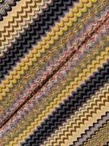 Capa tipo poncho de tejido de punto con diseño de zigzag multicolor en tonos amarillo, gris, negro y beige. Presenta cuello en V y flecos en el borde inferior.