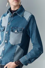 Campera recta de denim rígido con combinación de recortes en diferentes tonos de azul, cuello camisero, cierre frontal con botones metálicos, bolsillos con solapa y botones metálicos en el pecho, y puños con detalle de encaje negro.