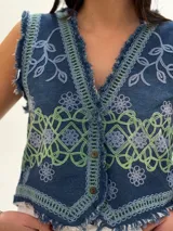 Chaleco corto de denim azul con bordados florales en blanco y marrón, detalle desflecado en escote, sisas y ruedo. Escote en V y cierre frontal con botones. Estilo boho chic.