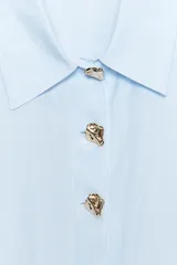 Camisa celeste a rayas verticales, de popelín, con cuello solapa, manga larga y cierre frontal con botones dorados.
