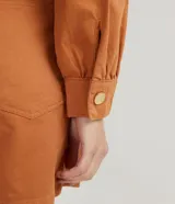 Mono corto color ladrillo, de sarga de algodón, con cuello mao, mangas abullonadas largas, cierre frontal con botones dorados, bolsillos laterales y cintura marcada.