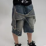 Bermuda de jean azul con efecto pre-lavado, tiro mediano y corte recto. Presenta un diseño deconstruido con parches de denim superpuestos y bordes deshilachados, incluyendo tiras de tela colgantes con botones.