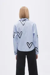 Camisa celeste a rayas verticales blancas, con cuello clásico, cierre frontal con botones y estampado de corazones negros.