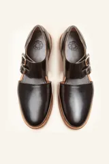 Zapato tipo monk strap con aberturas laterales, confeccionado en cuero vacuno color bordó. Presenta doble tira con hebillas metálicas plateadas en el empeine y suela de cuero con taco bajo.