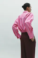 Camisa oversize rosada con rayas verticales blancas y celestes, cuello solapa, manga larga con puños abotonados, bolsillo de plastrón en el pecho y pinza en la espalda.