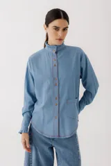 Camisa celeste de jean con cuello alto con volados y mangas largas abullonadas con puños con volados. Presenta botones de madera y costuras rojas a contraste.