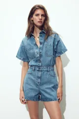 Mono corto de denim azul, con cuello solapa, manga corta, bolsillos delanteros y traseros, cintura elástica en la espalda y cierre frontal con cremallera y botones metálicos.