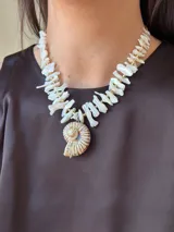 Collar corto con cuentas irregulares de nácar color blanco y dije de caracol con detalles dorados. Cierre con gancho y cadena de acero quirúrgico para regular el largo.