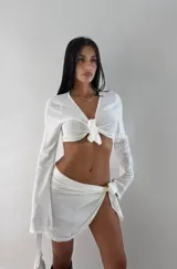 Conjunto de dos piezas color blanco, compuesto por un top manga larga anudado al frente y una minifalda pareo anudada lateralmente.