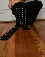 Pantalón negro acampanado de tiro bajo, con detalle de ojalillos metálicos en una pierna y faja desmontable.