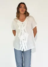 Camisa blanca de lino y algodón con cuello en V, mangas cortas y volados frontales. Cierre con botones.