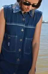 Vestido corto de jean gris con botones frontales, sin mangas, con cuello camisero y bolsillos en el pecho.