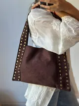 Bolso tipo tote de gamuza color marrón chocolate, con laterales y correa de hombro decorados con tachas doradas.