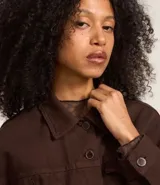 Campera marrón de sarga de algodón, corte regular, cuello inglés, manga larga, abotonada al frente, con bolsillos con solapa en el pecho y pliegues en la parte baja.