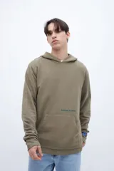 Canguro verde oliva de felpa liviana teñido en prenda, con capucha, bolsillo tipo canguro y puños acanalados. Estampa con la leyenda "UNFRM-STATE" en el pecho y detalle de estampa en la manga. Corte recto.