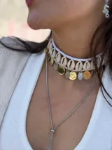 Gargantilla choker con hilo trenzado color blanco, adornada con conchas marinas tipo buzios y medallitas bañadas en oro y acero quirúrgico. Se cierra con cadena de acero quirúrgico y ganchito para ajustar el largo.