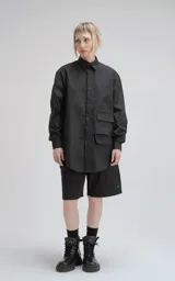 Camisa negra de manga larga, confeccionada en poplín de algodón. Presenta un calce oversize, cuello camisero, cierre frontal de botones y dos bolsillos cuadrados superpuestos en el lado derecho del pecho.