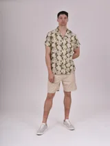 Camisa de manga corta con estampado de hojas color verde sobre fondo beige, cuello solapa y bolsillo en el pecho.