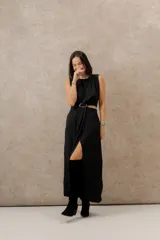 Vestido largo negro con tajo frontal y cinturón con hebilla dorada. Sin mangas y con cuello redondo.