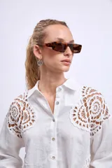 Camisa blanca de lino con cuello clásico, cierre frontal con botones y mangas largas con puños abotonados. Presenta apliques de encaje en hombros y bolsillos delanteros.