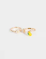 Set de dos anillos dorados con dijes: uno con forma de margarita blanca y otro con forma de limón amarillo y verde.