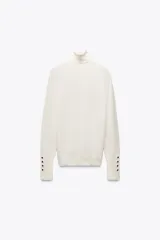 Jersey de punto color blanco, de cuello alto y manga larga con detalle de botones en el puño.