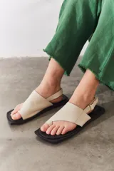 Sandalias de cuero color crema con tira ancha sobre los dedos, suela plana cuadrada color negro y tira trasera ajustable.