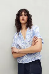 Camisa de manga corta con estampado de hojas blancas sobre fondo celeste, cuello solapa y botones de nácar al frente. Confeccionada en tejido liviano de sutil textura.