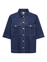 Camisa de denim azul con corte recto, mangas cortas anchas, cuello clásico, cierre frontal con botones metálicos y dos bolsillos de parche en el pecho.
