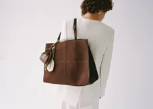 Bolso tipo shopper de piel de serraje color marrón, con diseño de paneles cosidos en el frente y bordes ribeteados en cuero liso oscuro. Posee dos asas de hombro de cuero liso oscuro y cierre de botón magnético.