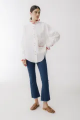 Camisa blanca de lino con cuello mao, manga larga abullonada y puños con detalle de crochet rojo. Tiene bolsillo de crochet en el pecho y cierre frontal con botones de madera.