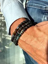 Set de tres pulseras elásticas con cuentas de piedra. Una de piedra volcánica negra con separadores plateados, otra de piedra ojo de tigre y piedra volcánica negra con separadores plateados, y otra de piedra volcánica negra con cuentas de piedra verde y separadores plateados.