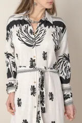 Vestido camisero midi de lino y viscosa color crudo con estampado de palmeras en negro. Tiene cuello camisero, mangas largas con puños y cinturón de la misma tela.