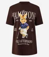 Remera de algodón color marrón oscuro con cuello redondo y manga corta. Presenta un estampado localizado con el texto "The Hamptons New York" y la ilustración de un conejo vestido de golfista.