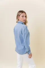 Camisa de denim celeste lavado, con cuello clásico y cierre de botones. Presenta bordado blanco en los bordes superiores de los bolsillos frontales y en los puños.