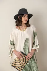 Vestido largo color natural con estampado de hojas de palmera en tonos verdes. Tiene corte amplio tipo túnica, cuello redondo, mangas tres cuartos, bolsillos laterales y ruedo con ribete.