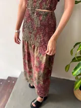 Conjunto de dos piezas compuesto por un top y una falda larga. Ambas prendas tienen un frunce elástico y un estampado floral en tonos verde oliva, rojo oscuro y negro. El top es de breteles finos y tiene espalda abierta regulable. La falda es larga y con cintura elastizada hasta la cadera.