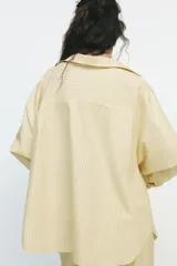 Camisa oversize a rayas amarillas y blancas, de cuello solapa y manga larga tipo murciélago acabada en puño con hombro caído. Tiene un bolsillo de plastrón en el delantero y cierre frontal con botones.