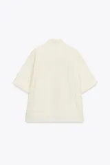 Camisa color crema de cuello solapa y manga corta con vuelta. Tiene un bolsillo de parche en el delantero, bajo asimétrico con aberturas laterales y cierre frontal con botones.