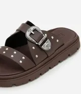 Sandalia plana negra con dos tiras anchas sobre el empeine, una con hebilla metálica estilo western y tachas plateadas.