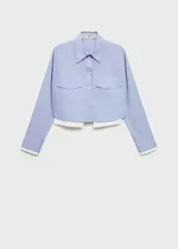 Camisa celeste de popelina de algodón, corte crop, con cuello camisero, cierre frontal con botones, dos bolsillos de solapa en la parte delantera y mangas largas con puños abotonados en color blanco.