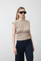 Top sin mangas color beige, confeccionado en tejido acanalado. Presenta hombreras definidas y un recorte fruncido bajo el busto que ajusta la silueta.