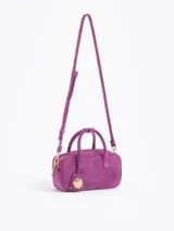 Cartera tipo baulito de cuero descarne vacuno en color fucsia, con diseño compacto y estructura rígida. Presenta doble asa de mano, logo de la marca grabado en relieve en el frente y un charm metálico en forma de vaquita de San Antonio colgando de una de las asas.