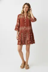 Vestido corto de poliéster, color terracota, con estampado tipo bandana en tonos beige. Presenta cuello en V con botones frontales y mangas 3/4 con volado en el puño.