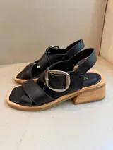 Sandalia de cuero vacuno color marrón, con tiras cruzadas en el empeine y hebilla metálica plateada. Posee taco de madera de 5 cm de altura y horma de punta cuadrada.