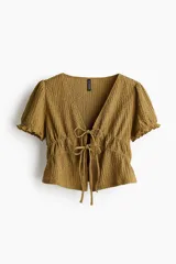 Top corto color mostaza con textura arrugada, escote en V profundo y mangas cortas abullonadas con elástico en el borde. Presenta un ajuste de cordón fruncido en la cintura que se ata con un lazo doble en el frente.