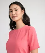 Blusa blanca de poliéster con cuello redondo, mangas cortas, cierre trasero en forma de gota, pequeñas aberturas laterales y acabados en pespuntes en las mangas y el cuello.